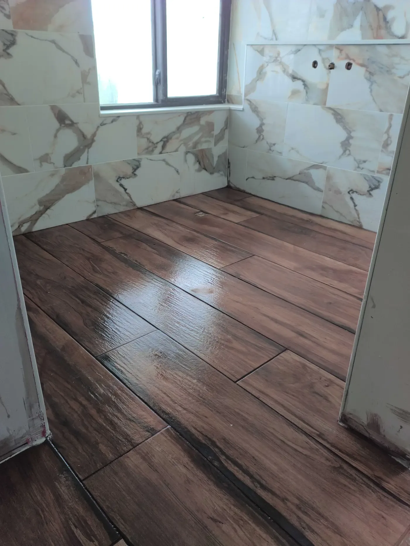 Bathroom renovation with dark wood-look plank floor tile, large marble-look wall tile around a window, and plumbing stubs on the shower wall