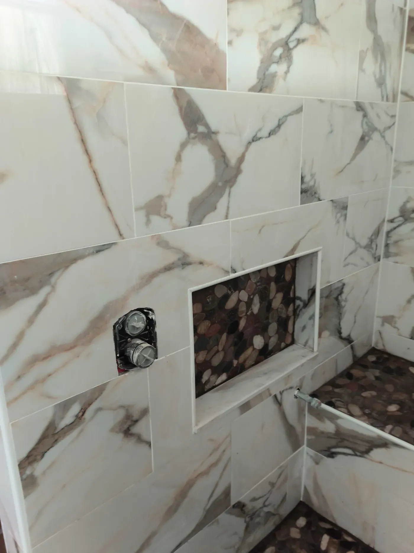 Custom shower niche lined with river rock pebble mosaic, framed in marble-look tile, with shower valve rough-in and lower outlet stub