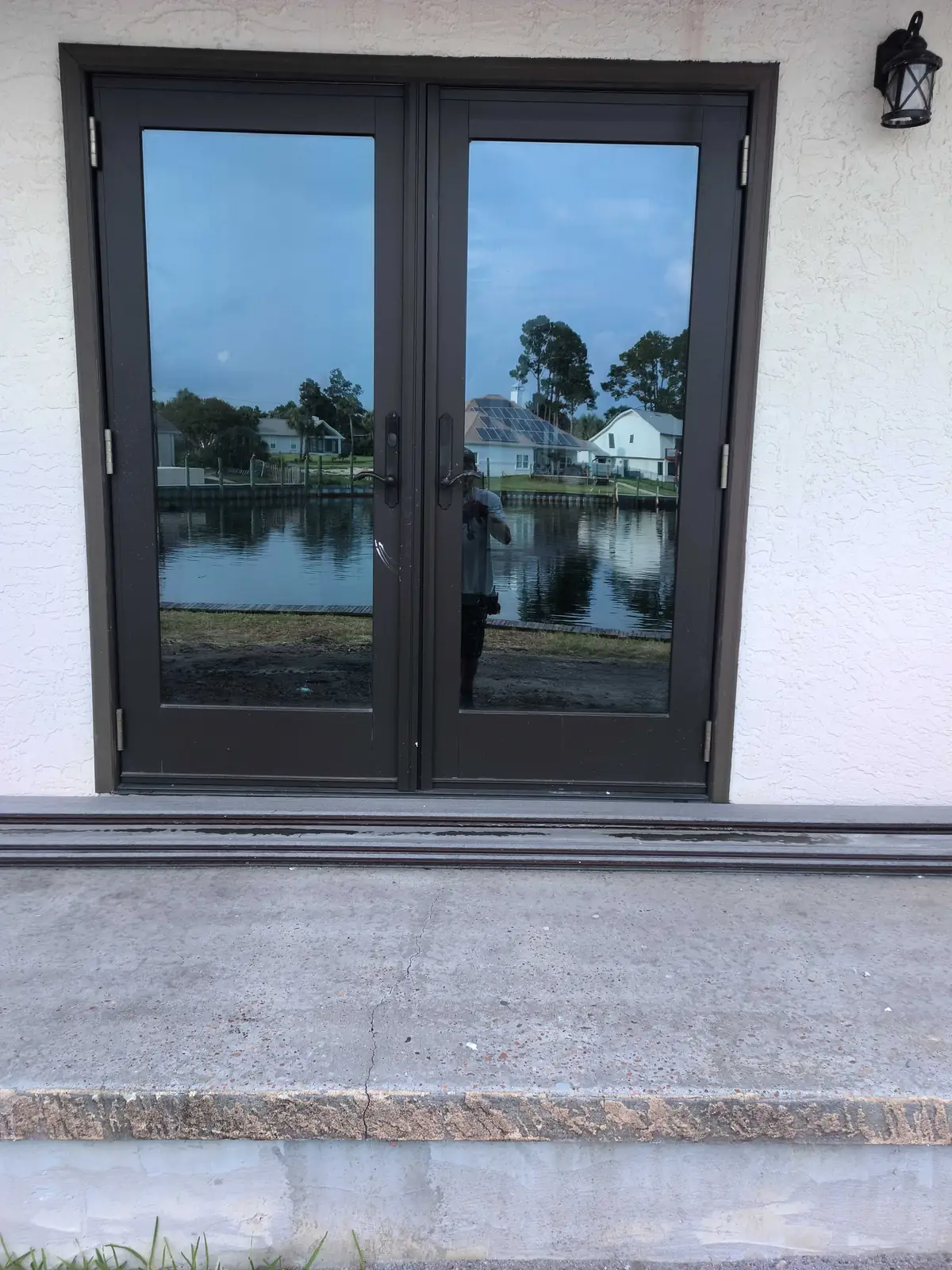 Dark-framed glass French doors in a white stucco wall with lantern light, threshold tracks, and a cracked concrete stoop on a Florida waterfront home