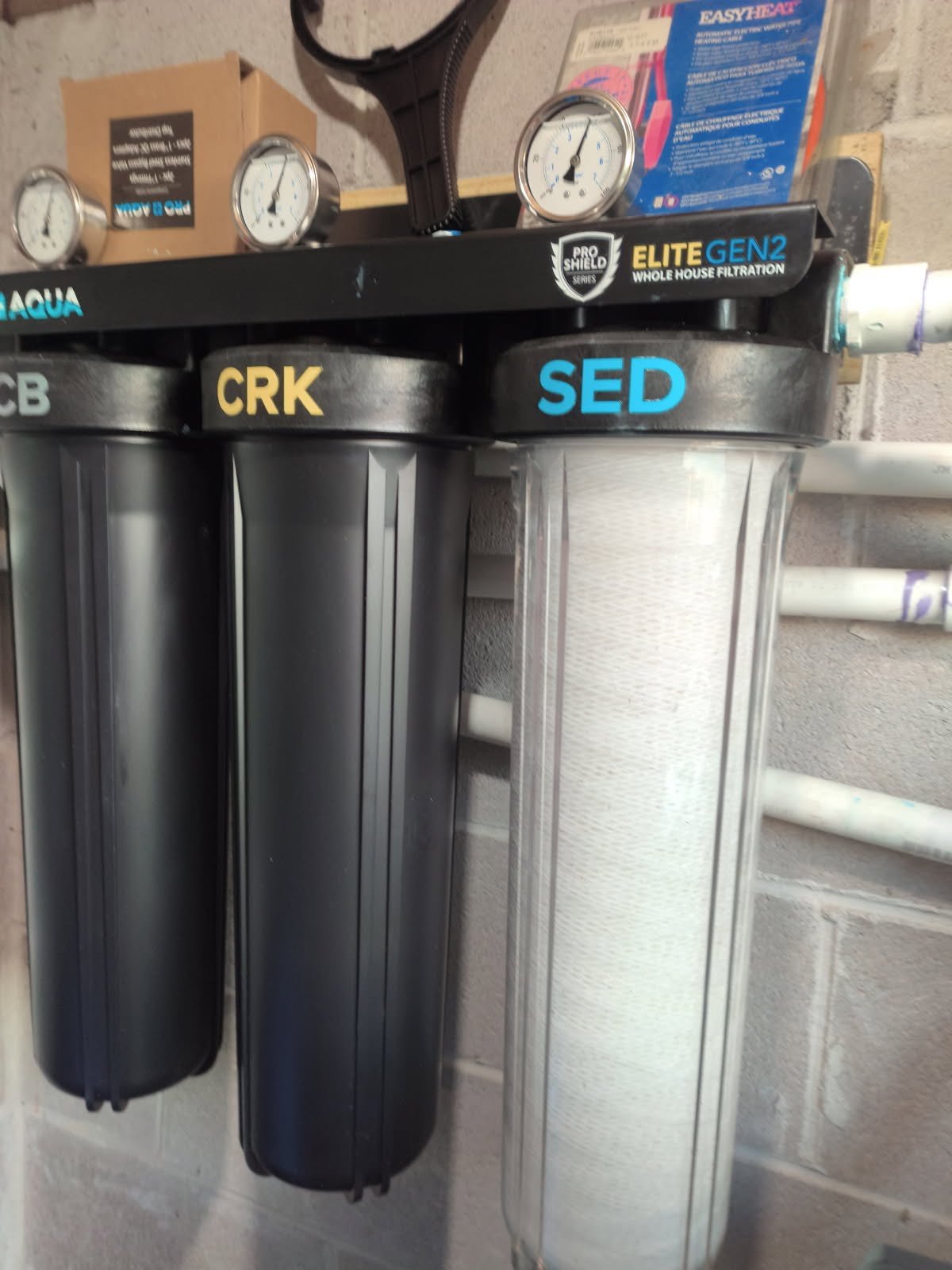 Pro Aqua Elite Gen2 whole house filtration system with SED sediment, CRK, and CB carbon block stages and pressure gauges mounted on a cinder block utility wall