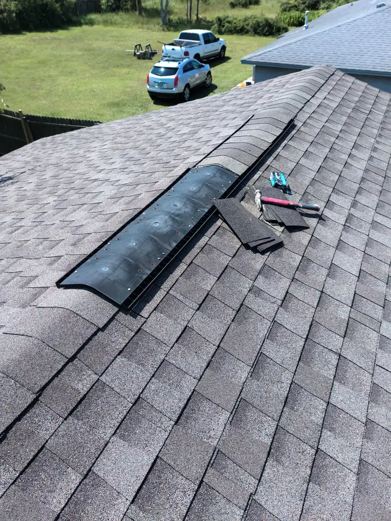 Ridge vent roofing repair in progress with black plastic vent nailed along the roof peak, cap shingles, hammer, and nail box on architectural shingles